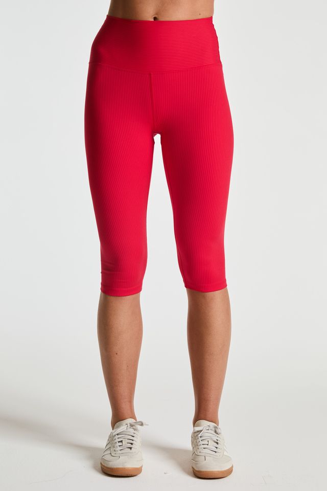 A model from the waist down wearing bright red ribbed capri leggings and white sneakers, standing against a plain white studio background.