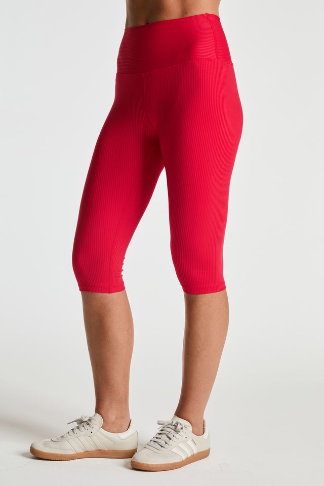 A model wearing bright red ribbed capri leggings and off-white sneakers, shown from the waist down against a light gray studio background.