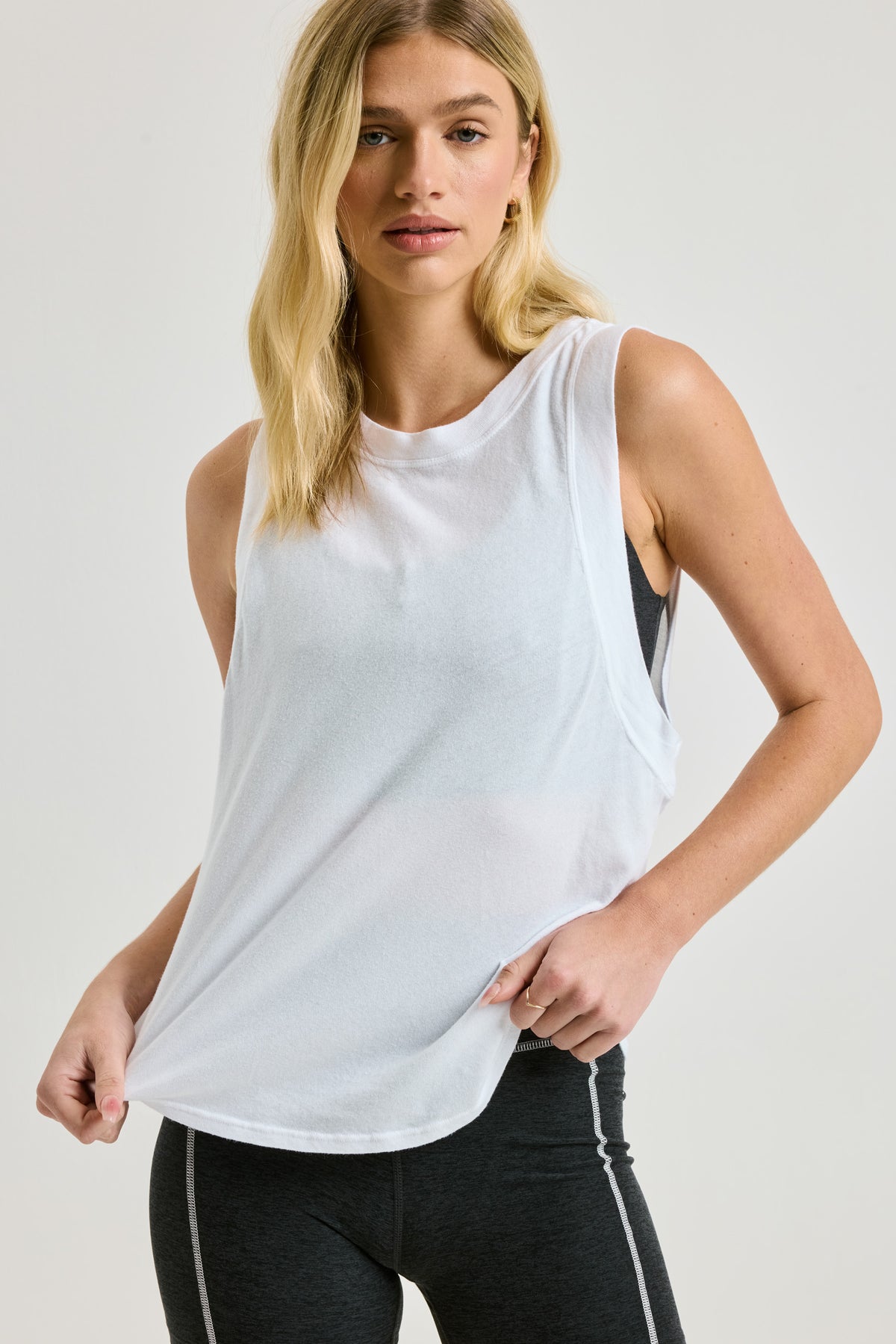 A blonde woman modeling a white sleeveless athletic tank top with black athletic leggings, photographed against a light gray studio background.