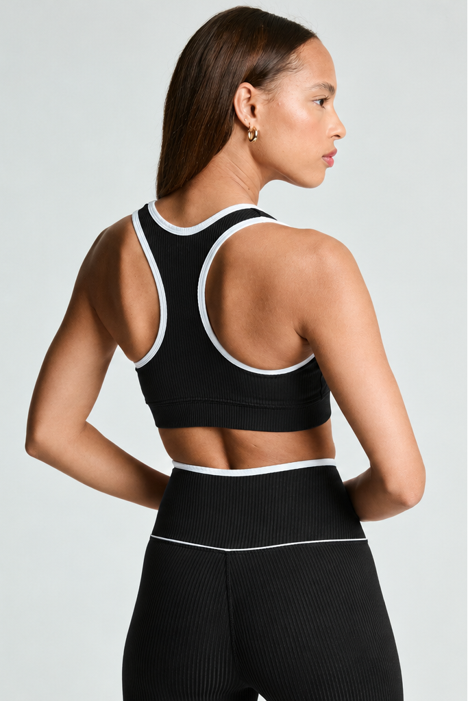 A woman shown from the back wearing a black ribbed sports bra and matching high-waisted leggings with white trim, against a light gray studio background.
