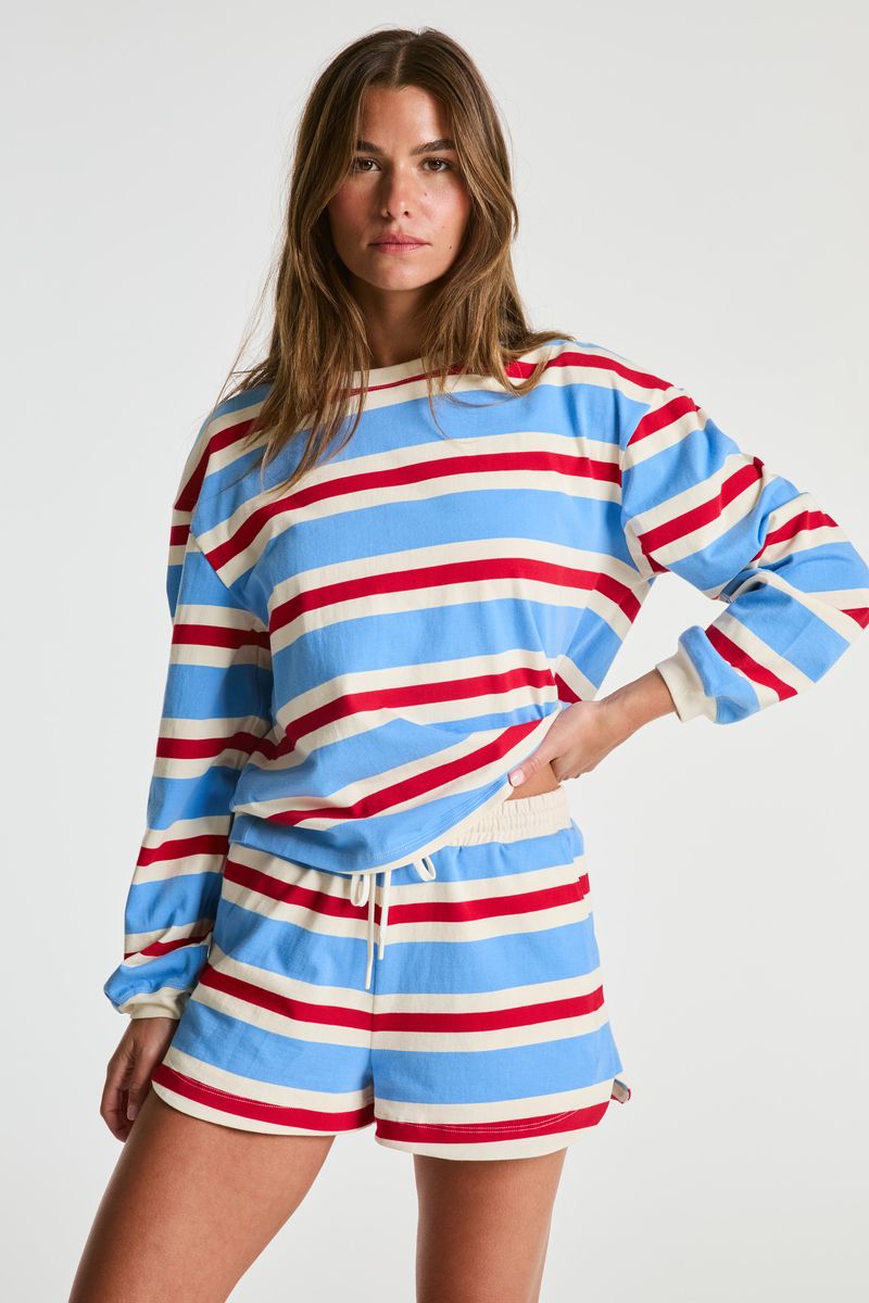 A woman modeling a matching lounge set with a long-sleeve crewneck sweatshirt and shorts, both featuring horizontal stripes in light blue, red, and off-white, against a plain light gray studio background.