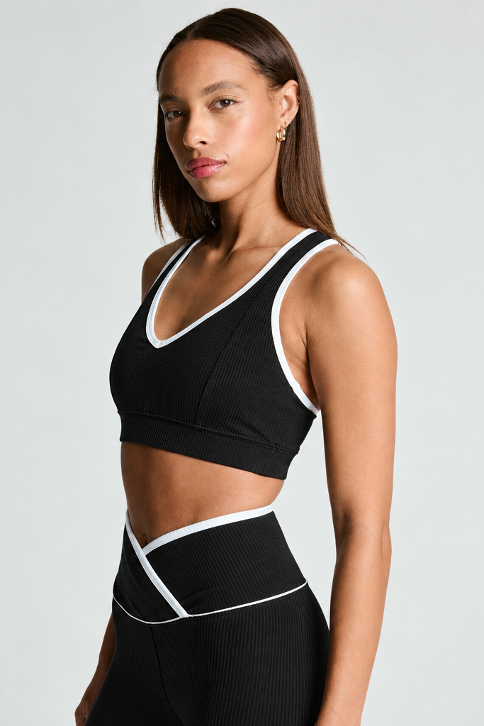 A woman modeling a black ribbed sports bra and matching high-waisted leggings with white contrast trim, photographed against a light gray studio background.